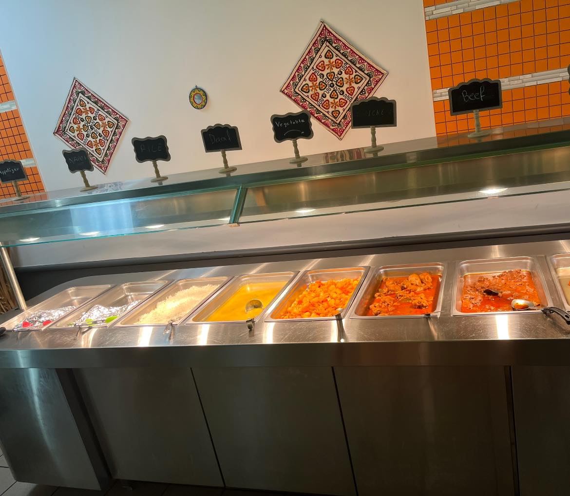 The Desi Meal serving counter with handwritten chalkboard signs for naan, rice, daal, vegetable, chicken and beef